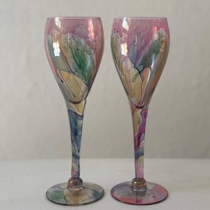 Könnte beinhalten: Zwei handbemalte Sektgläser. Die Gläser haben einen rosafarbenen Farbton mit farbenfrohen abstrakten Blumendesigns in Grün, Gelb und Lila. Die Stiele und Basen sind ebenfalls mit aufeinander abgestimmten Farben bemalt, was einen stimmigen und künstlerischen Look erzeugt.