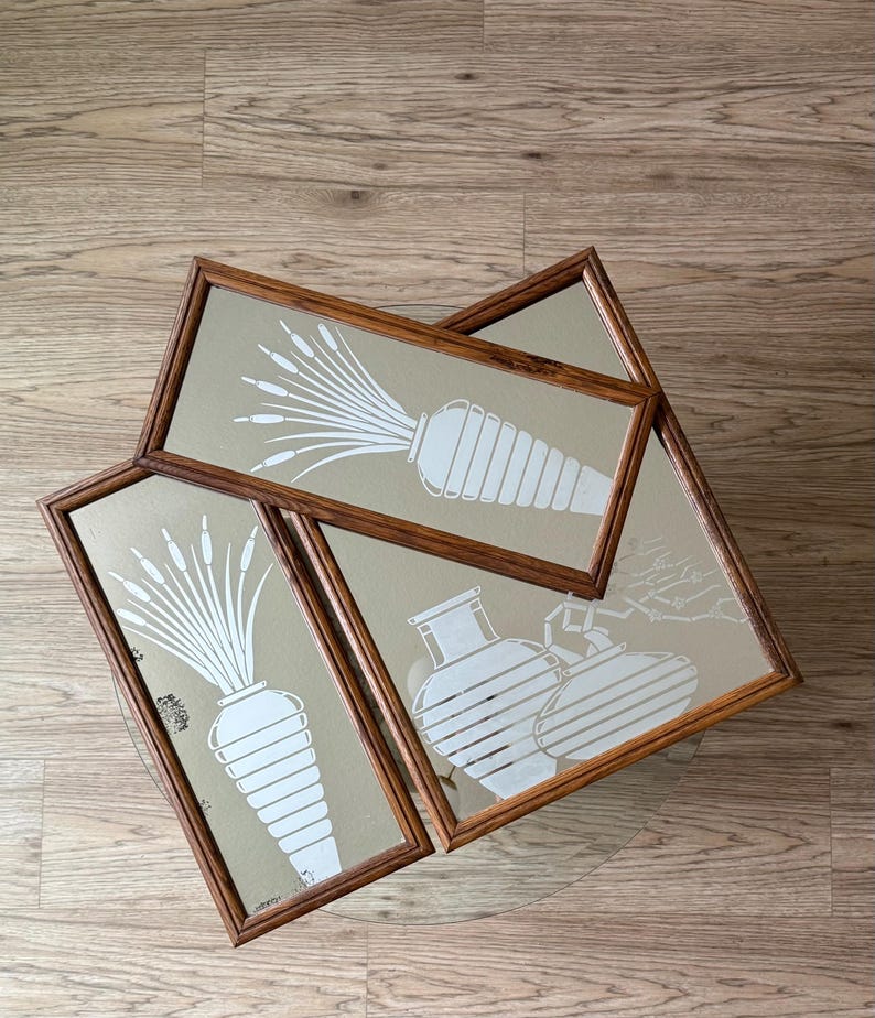 May include: Three framed wall mirrors with a white silhouette design of stylized plants and vases. The mirrors have a brown wooden frame.