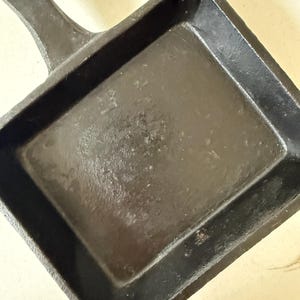 Puede incluir: Una sart&eacute;n cuadrada de hierro fundido negro con un asa. La sart&eacute;n tiene una superficie de cocci&oacute;n rectangular y laterales elevados. El asa tiene un agujero para colgar. La sart&eacute;n es de hierro fundido.