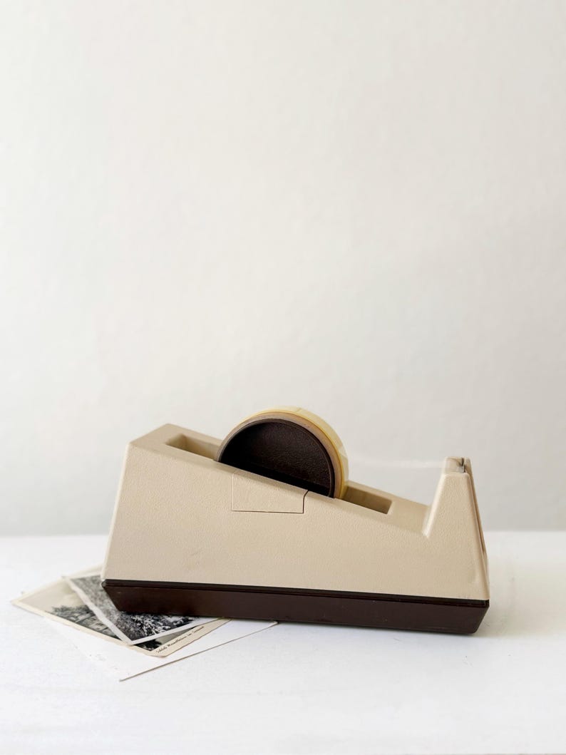 Puede incluir: Un dispensador de cinta adhesiva beige y marr&oacute;n vintage con un rollo de cinta. El dispensador tiene forma rectangular con una base marr&oacute;n y una parte superior beige. Unas cuantas fotograf&iacute;as en blanco y negro est&aacute;n delante del dispensador.
