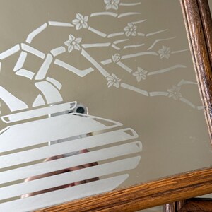 May include: A wooden framed mirror with an etched design of a vase with a flowering branch. The design is in white on a mirrored surface.