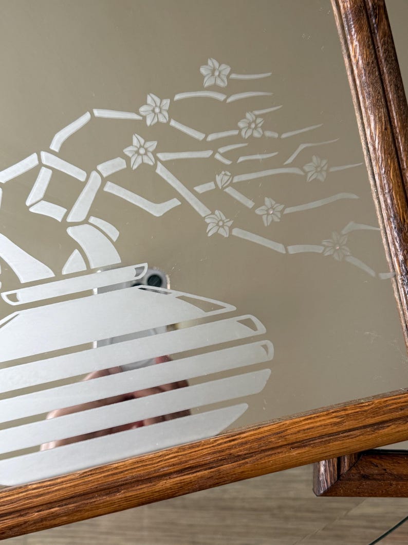 May include: A wooden framed mirror with an etched design of a vase with a flowering branch. The design is in white on a mirrored surface.