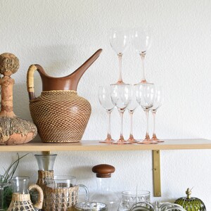 May include: A wooden shelf with various decorative items, including a brown woven pitcher, clear glass wine glasses, and a clear glass bottle with a woven cover. There are also two white ceramic birds, a green glass jar, and a green air plant.