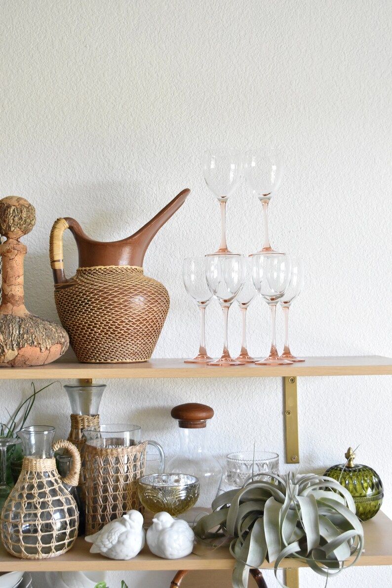 May include: A wooden shelf with various decorative items, including a brown woven pitcher, clear glass wine glasses, and a clear glass bottle with a woven cover. There are also two white ceramic birds, a green glass jar, and a green air plant.
