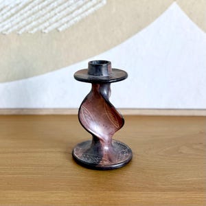 May include: A dark brown and reddish-brown candlestick holder with a twisted design. The candlestick has a circular base and a flared top, with etched leaf patterns. The holder is made of a dark material, possibly stone or wood.