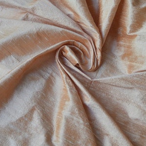 May include: A close-up of a peach-coloured silk fabric with a subtle shimmer. The fabric is draped and folded, creating soft, flowing lines.