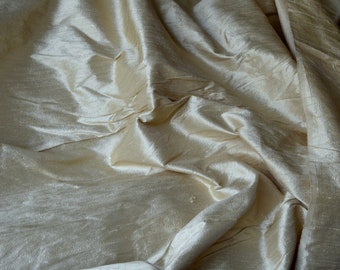 May include: A close-up of a smooth, ivory-colored fabric with a subtle sheen. The fabric is draped and folded, creating soft, flowing lines.