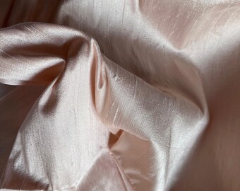 May include: A close-up of a soft, pale pink fabric with a subtle sheen. The fabric is draped and folded, creating a smooth, flowing texture.