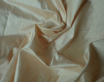 May include: A close-up of a smooth, pale peach-colored fabric with a subtle sheen. The fabric is draped and folded, creating soft, flowing lines.