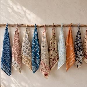 Pode incluir: Dez panos de tecido quadrados com vários padrões estampados em blocos em tons de azul, vermelho, laranja e creme, cada um atado no topo e pendurado num ramo de madeira horizontal. Os designs incluem formas geométricas, motivos florais e riscas.