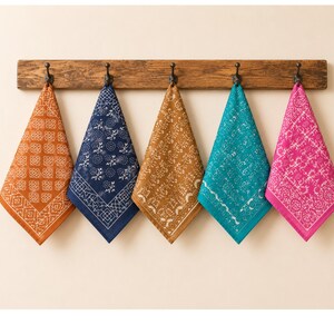 Puede incluir: Cinco pañuelos coloridos, naranja, azul marino, beige, turquesa y rosa, cuelgan de un soporte de madera. Cada pañuelo presenta un patrón blanco único. Los pañuelos están doblados en triángulos y colgados de ganchos negros.