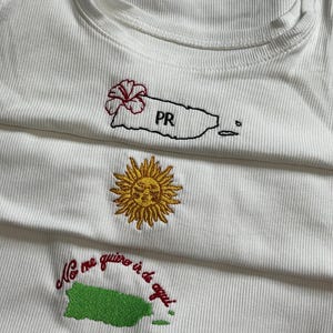 May include: White ribbed vest tops with embroidered designs. One features a red flower and the letters "PR" over a map outline. Another has a gold sun, and the third has the text "No me quiero ir de aqui" over a green map outline.