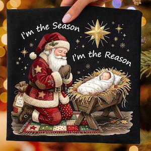 May include: Black graphic tee featuring Santa Claus kneeling in prayer, with a baby in a manger. The text reads "I'm the Season, I'm the Reason." A gold star and other stars are also present. A small bag with the text "Gift of Faith" is also present.