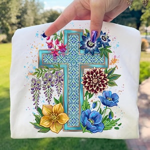 Puede incluir: Camiseta blanca con un diseño de cruz con borde turquesa y arreglos florales. Las flores incluyen flores azules, amarillas, moradas y granates, con hojas verdes y salpicaduras de pintura coloridas.