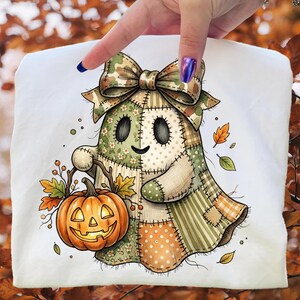 May include: A white t-shirt featuring a patchwork ghost design with a camouflage bow. The ghost holds a carved pumpkin with autumn leaves. The design incorporates various patterns and colors, including green, orange, and beige, creating a fall-themed aesthetic.
