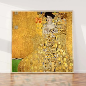 May include: A framed print of a Gustav Klimt painting. The artwork features a woman in a long, ornate gold dress with intricate patterns. The background is a rich gold, and the frame is a light wood color.