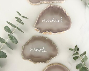 May include: Four agate stone place cards with names written in white script: "andrew", "michael", "nicole", and "elise". The stones are a light grey-brown color with a gold-colored edge. Eucalyptus sprigs are placed around the cards.