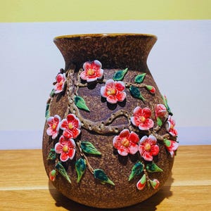 Könnte beinhalten: Eine braune Keramikvase mit strukturierter Oberfläche, verziert mit erhabenen roten und weißen Blüten, grünen Blättern und einem braunen Rankenmuster. Die Vase hat einen breiten, abgerundeten Körper und einen ausgestellten Rand.