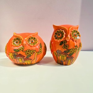 May include: Two orange ceramic owl figurines. The owls have large, detailed eyes and are decorated with yellow and green floral and house motifs. One owl is round, the other is upright.