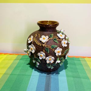 May include: A brown ceramic vase with a dark brown interior and a decorative floral design. The vase features white flowers with yellow centers and green leaves, adding a touch of nature-inspired elegance to any room. The vase is approximately 8 inches tall.