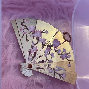 May include: A gold-colored, folding fan with a floral design in pink and brown. The fan is open, revealing the detailed artwork of cherry blossoms. The fan has a small ring at the top and a decorative rivet at the base.