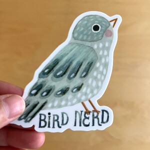 Pu&ograve; includere: Un adesivo con un'illustrazione ad acquerello di un uccello nei toni del grigio e del blu. L'uccello ha una guancia rosa e zampe marroni. Il testo "BIRD NERD" &egrave; sotto l'uccello in un carattere blu scuro.