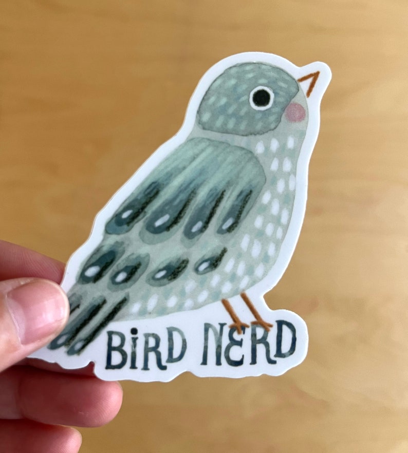 Pu&ograve; includere: Un adesivo con un'illustrazione ad acquerello di un uccello nei toni del grigio e del blu. L'uccello ha una guancia rosa e zampe marroni. Il testo "BIRD NERD" &egrave; sotto l'uccello in un carattere blu scuro.
