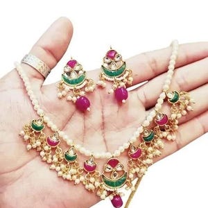 May include: A gold-toned necklace and earring set featuring a pearl strand and ornate pendants. The pendants have a red and green design with small white beads and a dangling magenta bead. The earrings mirror the pendant design.