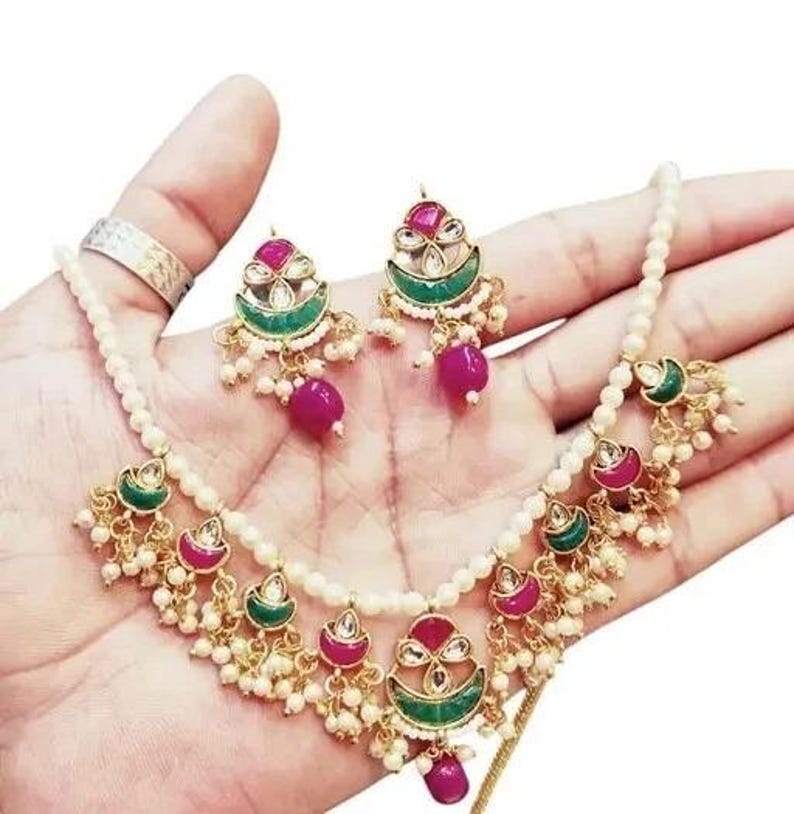 May include: A gold-toned necklace and earring set featuring a pearl strand and ornate pendants. The pendants have a red and green design with small white beads and a dangling magenta bead. The earrings mirror the pendant design.