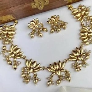 May include: A gold-colored jewelry set featuring a necklace and matching earrings. The necklace and earrings are adorned with lotus flower pendants and small, dangling beads. The set is displayed on a white surface.