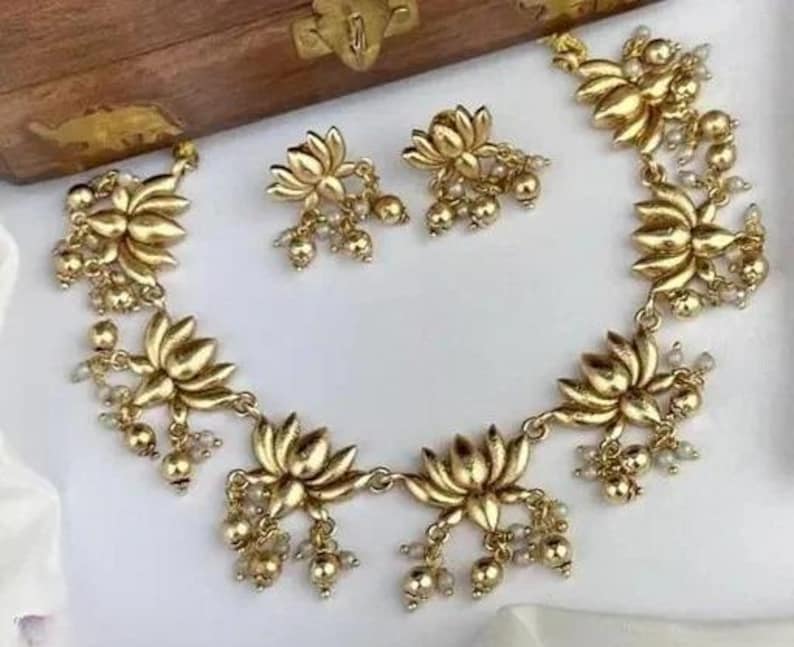 May include: A gold-colored jewelry set featuring a necklace and matching earrings. The necklace and earrings are adorned with lotus flower pendants and small, dangling beads. The set is displayed on a white surface.