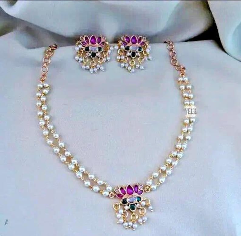 May include: A gold-toned necklace and matching earrings set. The necklace features a double strand of small, white pearls and a pendant with pink, green, and black gemstones. The earrings have a similar design, with a floral shape and dangling pearls.