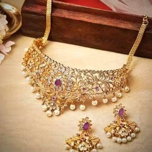 Puede incluir: Un conjunto de collar y pendientes dorados. El collar presenta un diseño ornamentado con piedras preciosas claras y moradas, y perlas blancas. Los pendientes reflejan el diseño del collar. Las joyas se exhiben sobre una superficie clara.