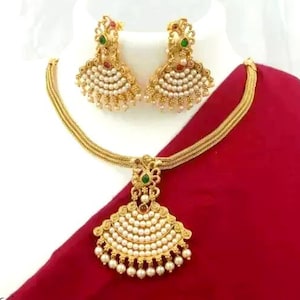 May include: Gold-toned jewelry set featuring a necklace and matching earrings. The necklace has a pendant with a fan-shaped design, adorned with white pearls and green and red accents. The earrings mirror the pendant's design. The set is displayed on a white bust against a red backdrop.