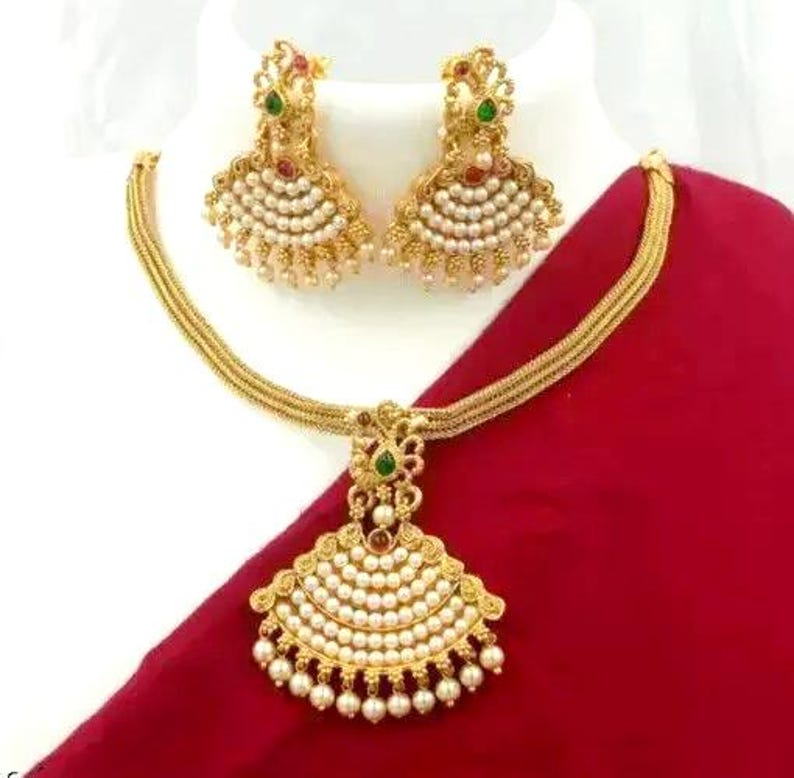 May include: Gold-toned jewelry set featuring a necklace and matching earrings. The necklace has a pendant with a fan-shaped design, adorned with white pearls and green and red accents. The earrings mirror the pendant's design. The set is displayed on a white bust against a red backdrop.
