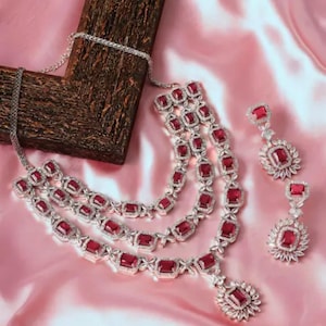 Puede incluir: Un conjunto de collar de tres hilos y pendientes a juego. La joyería presenta metal plateado con piedras rojas de corte cuadrado. Los pendientes tienen un diseño floral alrededor de la piedra roja. Se muestra sobre satén rosa.