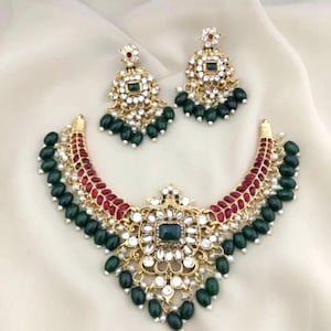 May include: A gold-toned necklace and earring set. The necklace features a central square green stone surrounded by white gems and a red and gold curved design. The earrings mirror the necklace design, with green bead drops.