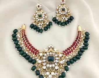 May include: A gold-toned necklace and earring set. The necklace features a central square green stone surrounded by white gems and a red and gold curved design. The earrings mirror the necklace design, with green bead drops.