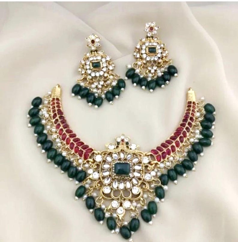 May include: A gold-toned necklace and earring set. The necklace features a central square green stone surrounded by white gems and a red and gold curved design. The earrings mirror the necklace design, with green bead drops.