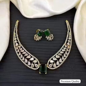 Peut inclure: Un ensemble collier et boucles d'oreilles doré avec des pierres précieuses claires et vert émeraude. Le collier a un design incurvé avec une grande pierre rectangulaire vert émeraude au centre. Les boucles d'oreilles sont de petits clous avec des pierres vert émeraude. Le texte "Premium Quality" est visible.