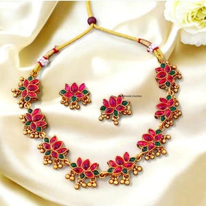 May include: A gold-toned necklace and earring set featuring red and green floral designs. The necklace and earrings have a flower-like design with red petals and green leaves. The set is displayed on a white satin surface.