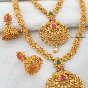 May include: Gold-toned jewelry set featuring two necklaces and matching earrings. The necklaces have intricate floral and leaf patterns, with pendants adorned with red and green gemstones. The earrings are bell-shaped with a red gemstone accent.
