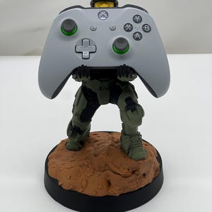 May include: A Master Chief figurine standing on a textured brown rocky base, holding a light gray Xbox wireless controller. The controller features a central Xbox logo button, a directional pad, two analog sticks with green accents, and A, B, X, Y buttons.