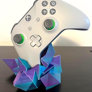 May include: A white Xbox controller rests on a geometric stand. The controller has gray buttons and green accents. The stand is a cluster of iridescent, multi-faceted shapes in shades of blue, purple, and teal.