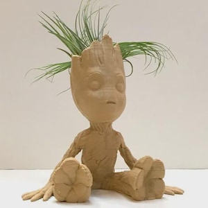 May include: A tan-colored Groot-shaped planter holding a green plant. The planter has a detailed, textured surface and is sitting with its legs extended. The plant has long, thin green leaves. The planter is a decorative item.