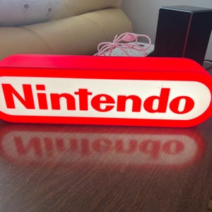 以下が含まれることがあります： 赤と白の任天堂ロゴのライトボックス。長方形のライトボックスは赤い縁取りがあり、白い楕円形の背景に「Nintendo」の文字が白で書かれています。 ライトボックスは茶色の表面にあります。