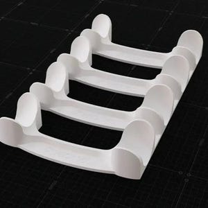 May include: A white plastic organizer rack featuring four parallel curved slots designed to hold items horizontally. The structure consists of a solid base with raised, rounded dividers at each end of the four slots, creating a uniform grid.