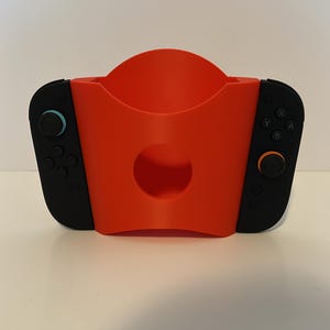 May include: A bright orange Nintendo Switch controller holder with a circular cutout. The holder has two black controllers attached on either side. The left controller has a blue joystick, and the right controller has an orange joystick.
