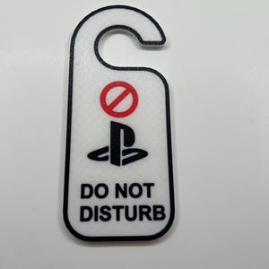 May include: White door hanger with a black border, featuring a red no-entry symbol above a black PlayStation logo. The words "DO NOT DISTURB" are printed in black below the logo.