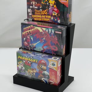 May include: A black display stand holding three video game boxes. The boxes are for Super Mario RPG, Super Metroid, and Mario Kart 64. Each box is in a clear protective case. The Nintendo logo is visible on each box.